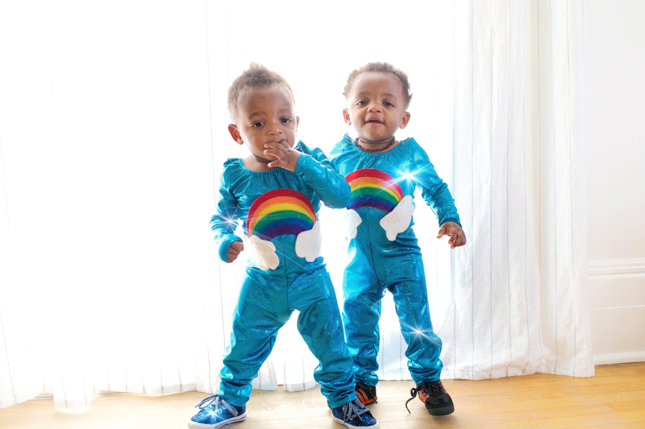 Two young children dressed in matching blue outfits with rainbow and cloud designs, standing on a wooden floor in front of sheer white curtains, with light shining through.