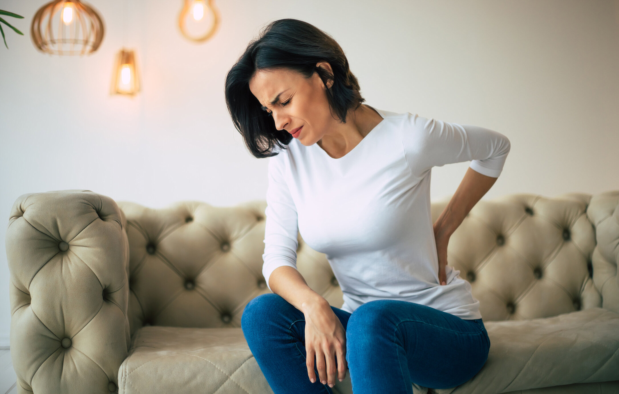 Woman dressed in a white shirt and blue jeans sitting on a beige tufted sofa, holding her lower back in pain, with a distressed expression.