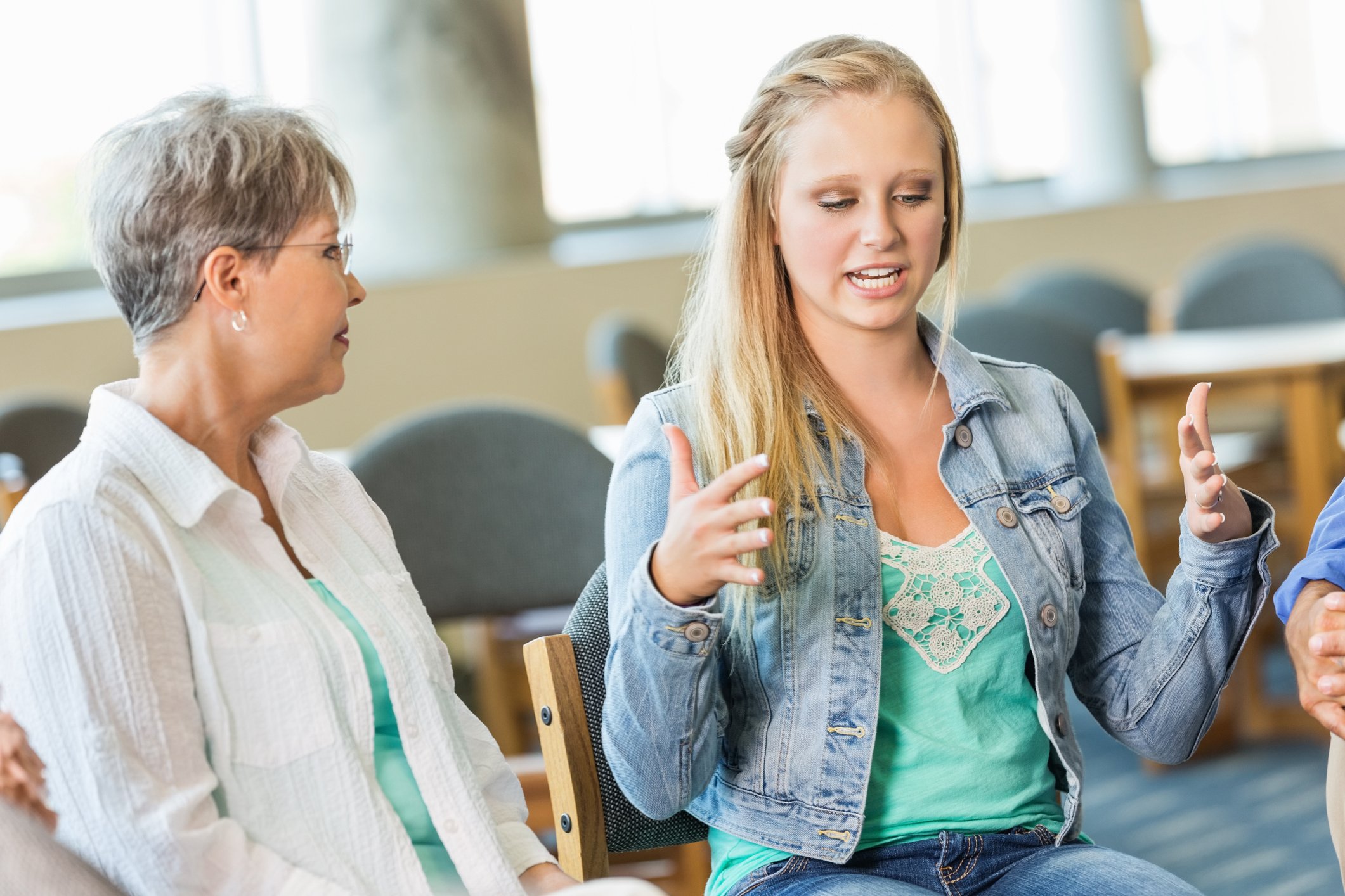 A young woman with long blonde hair, wearing a denim jacket, talks animatedly with a middle-aged woman with short gray hair, wearing glasses and a white blouse, in a room with large windows and empty chairs in the background.