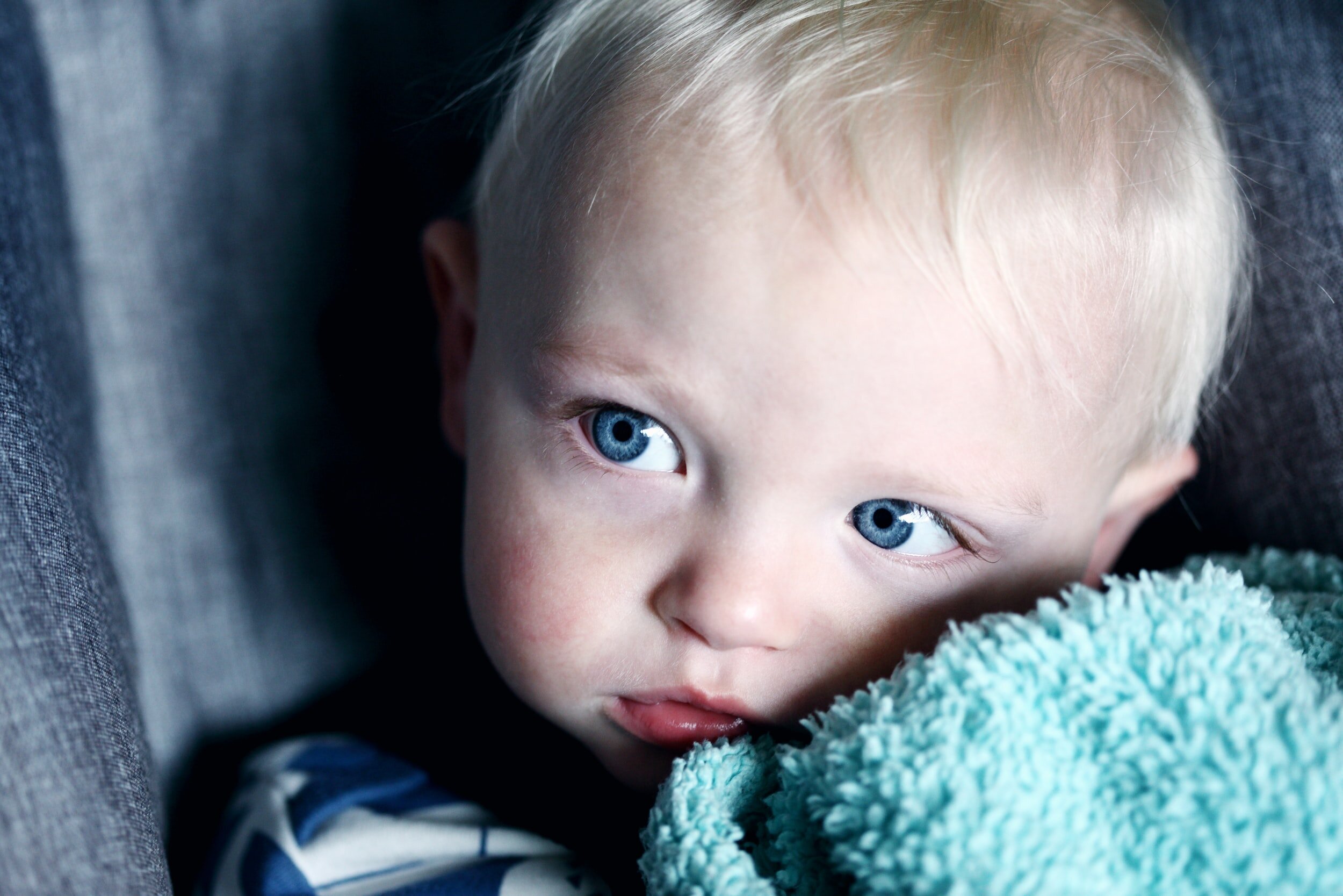 Close-up of a young blonde-haired boy with blue eyes resting his head on a soft, teal-colored blanket, gazing to the side, with a dark background.