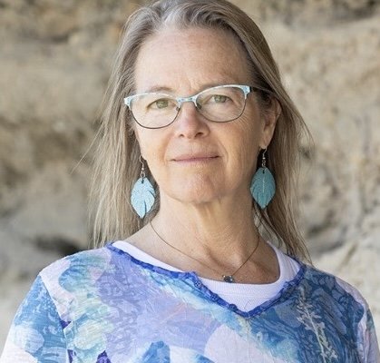 A woman with gray hair and glasses standing outdoors with a rocky background, wearing a blue patterned top and matching earrings.