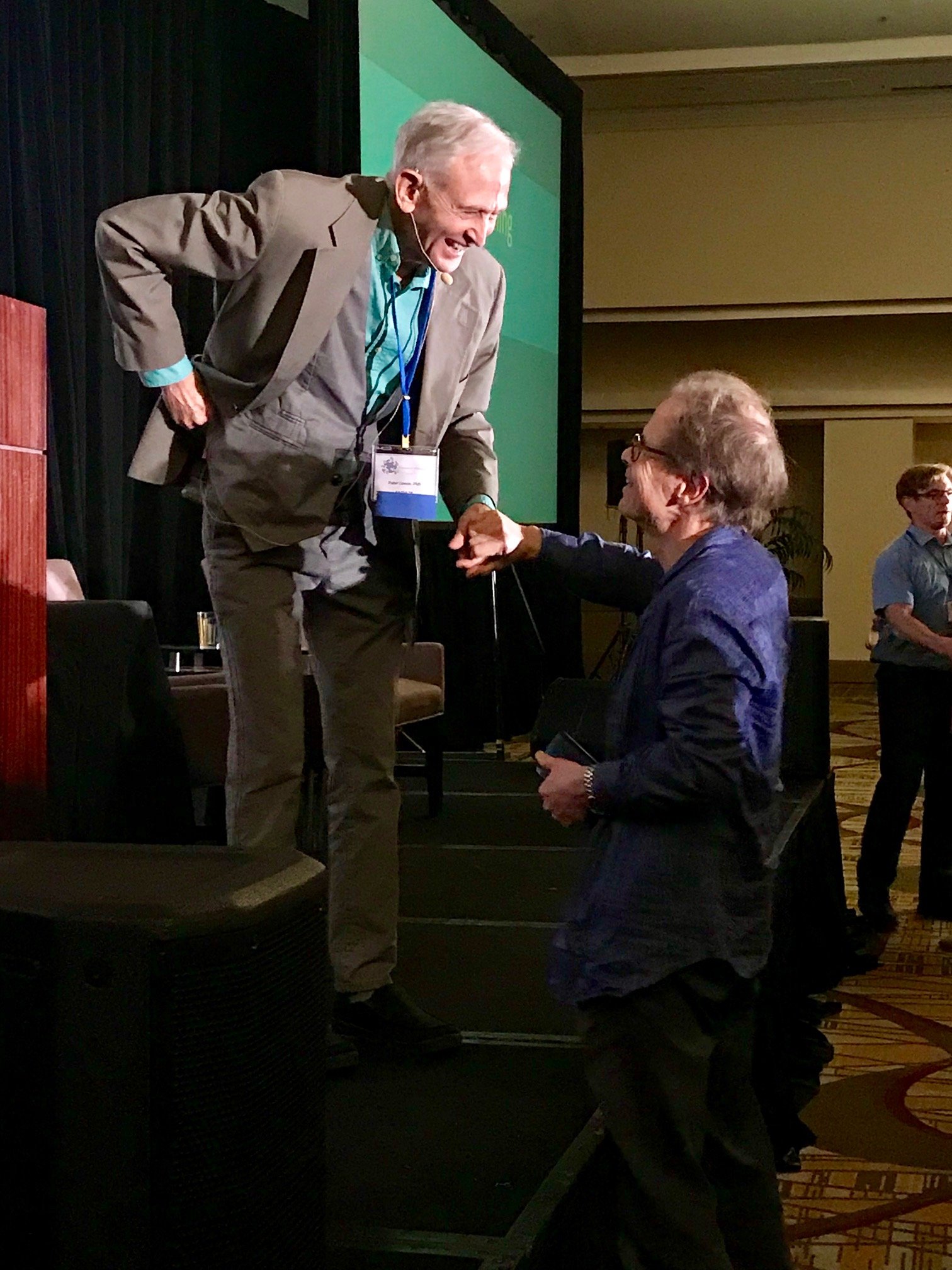 Two men shaking hands at a conference, one standing on a stage and wearing a gray suit, the other kneeling in front wearing glasses and a dark blazer.