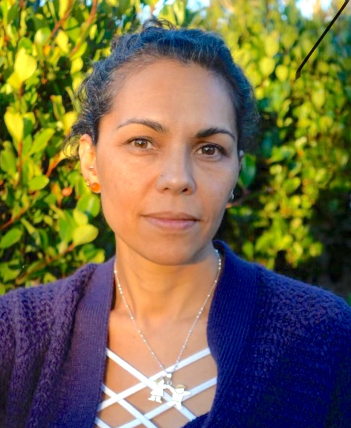 Person with dark hair wearing a purple sweater and necklace, standing in front of green foliage.